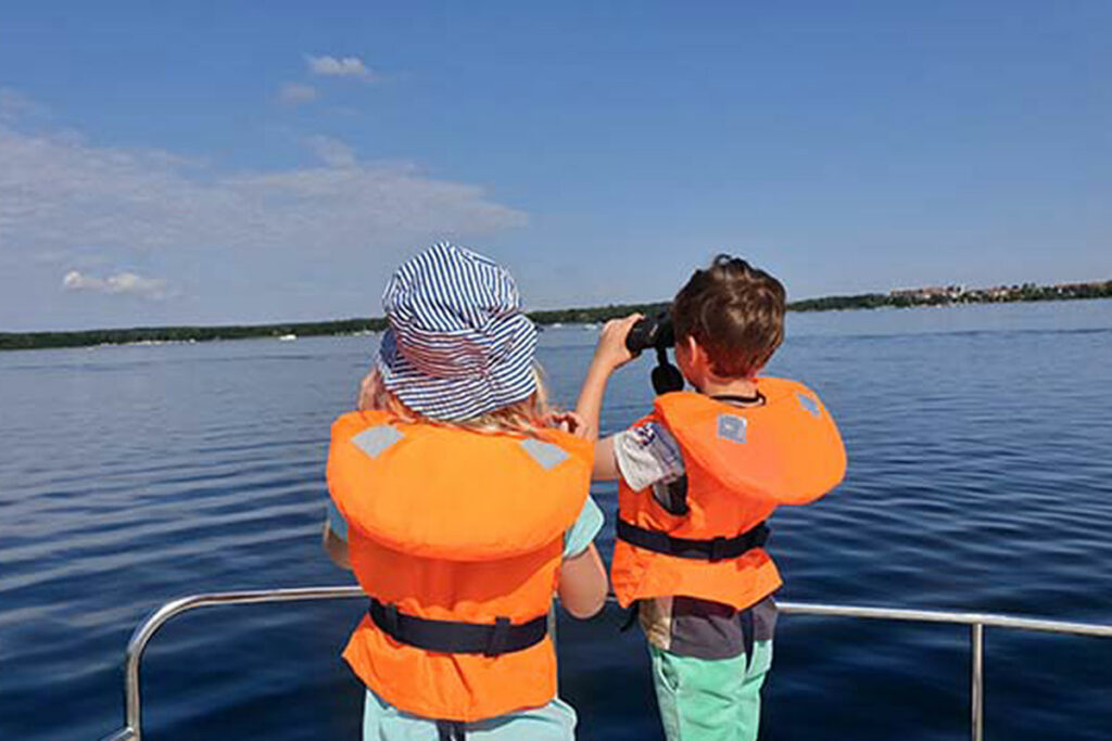 Zwei Kinder schauen mit einem Fernglas von Bord eines Hausbootes über der den See