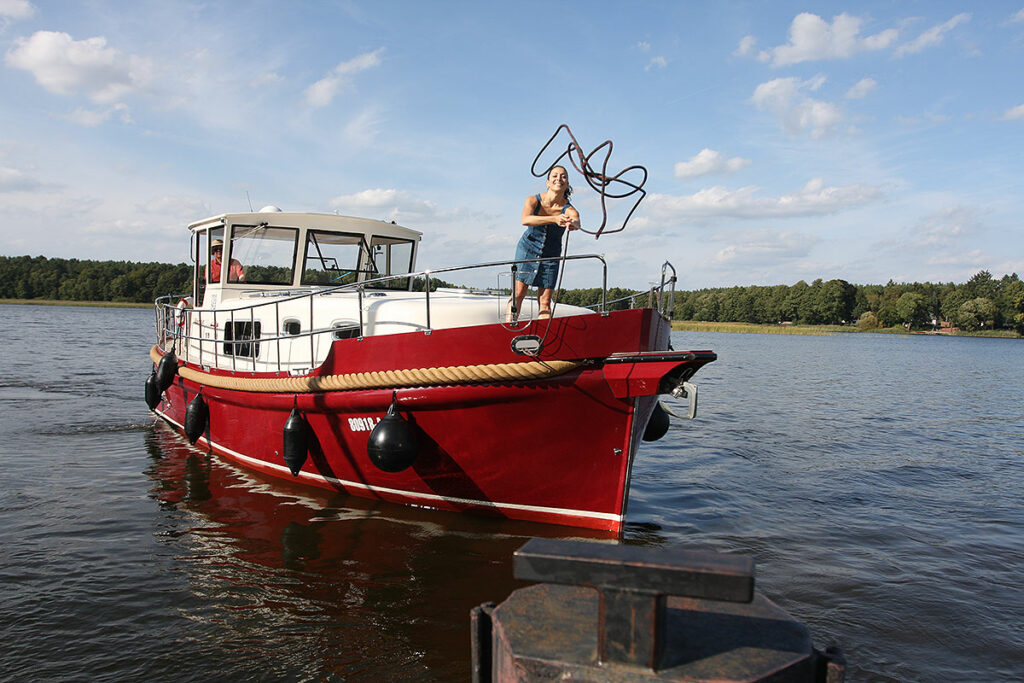 Eine junge Frau auf einem Hausboot vom Typ Riverboat wirft eine Leine über einen Poller zum Anlegen in der Marina am Röblinsee
