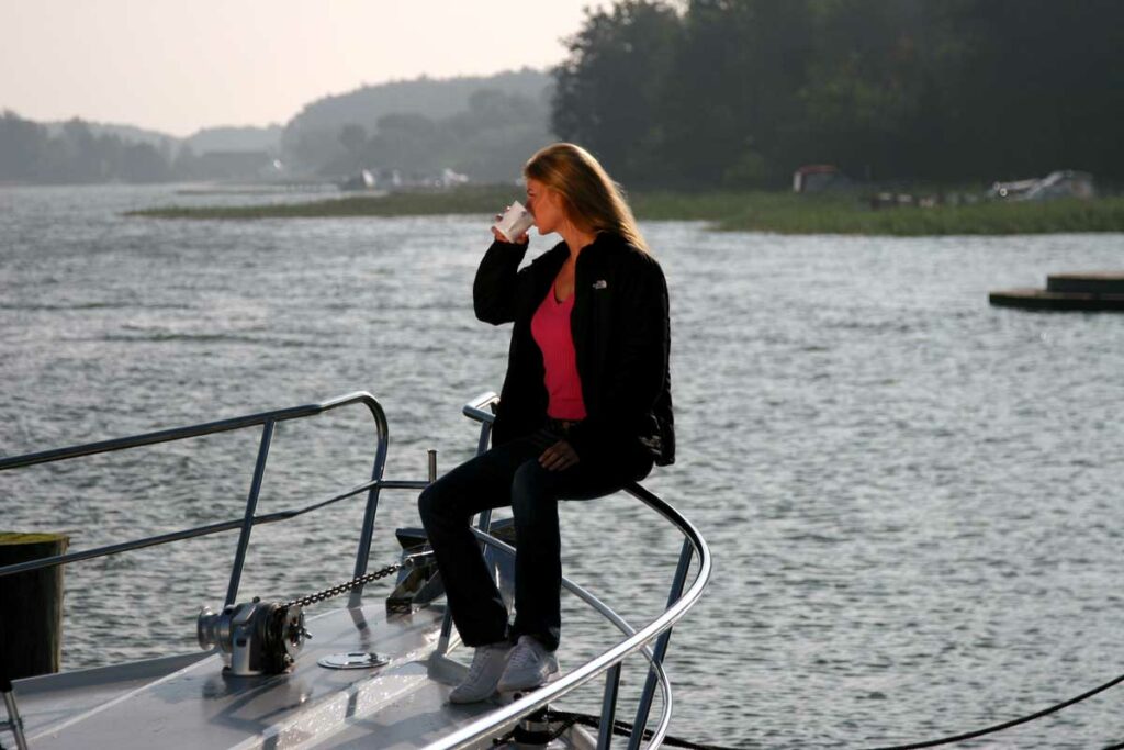 Frau sitzt entspannt im Bugkorb eines Motorbootes und genießt den Sonnenaufgang mit einem Kaffee in der Hand